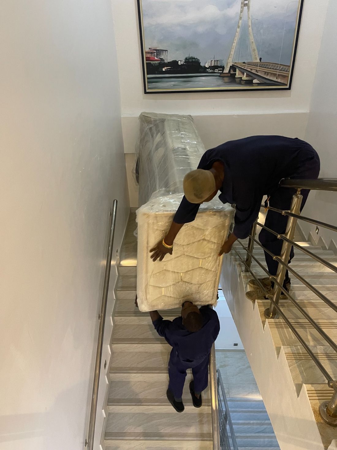 Two Pack n Moove movers in navy uniforms carrying a shrink-wrapped mattress down a staircase during a house move in Lagos
