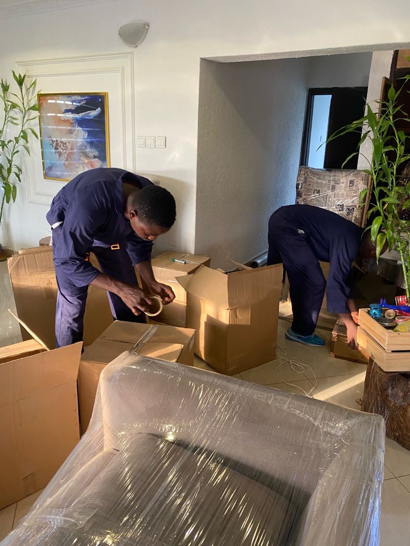Pack n Moove crew taping and sealing packed cartons in a Lagos living room beside a shrink-wrapped sofa