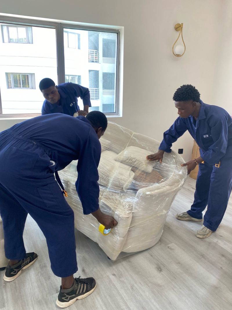 Three Pack n Moove movers shrink-wrapping a cream armchair to protect it before transport during an apartment move in Lagos